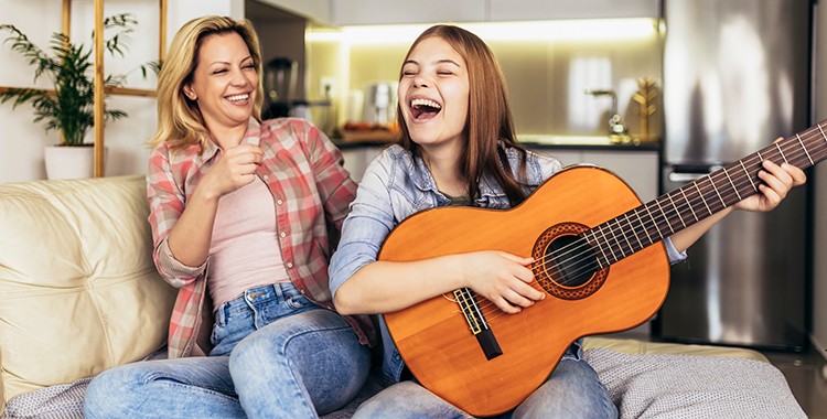 Uma fotografia de uma mãe rindo com a sua filha tocando violão. 