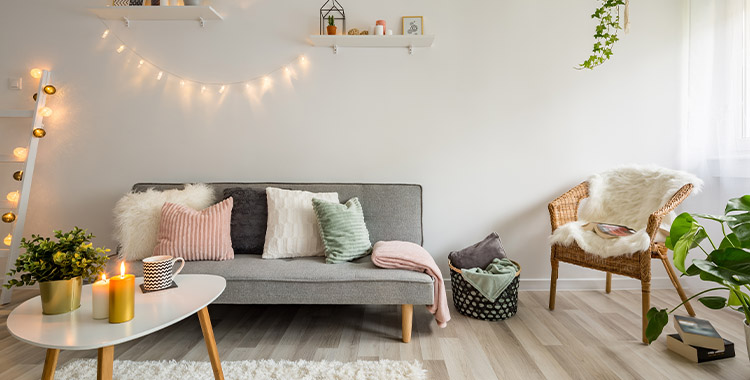 Uma fotografia que mostra uma sala com sofá pequeno, mesinha de cento em cima de um tapete quadrado branco, parede branca com luzes penduradas e plantas