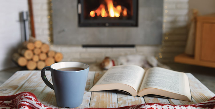 Uma fotografia de uma mesa com um livro aberto, uma xícara de café e ao fundo uma lareira e lenha do lado esquerdo