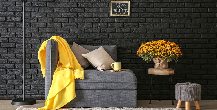 Uma fotografia de uma sala com parede de tijolinhos preta com um sofá cinza por sobre um tapete e um lençol amarelo 
