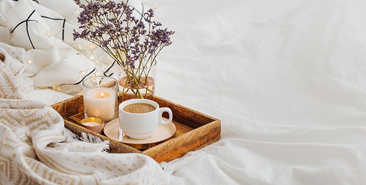 uma Fotografia de um café na cama com bandeja, xícara, vela e flores