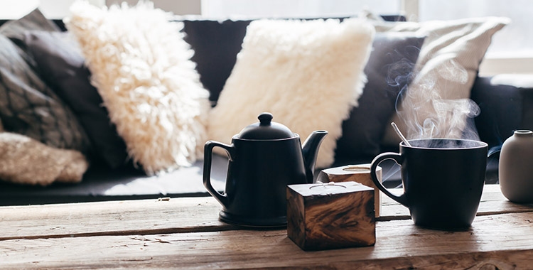 Uma fotografia focando uma mesa de madeira com um bule pequeno preto e uma xícara de café preto com almofadas no sofá ao fundo