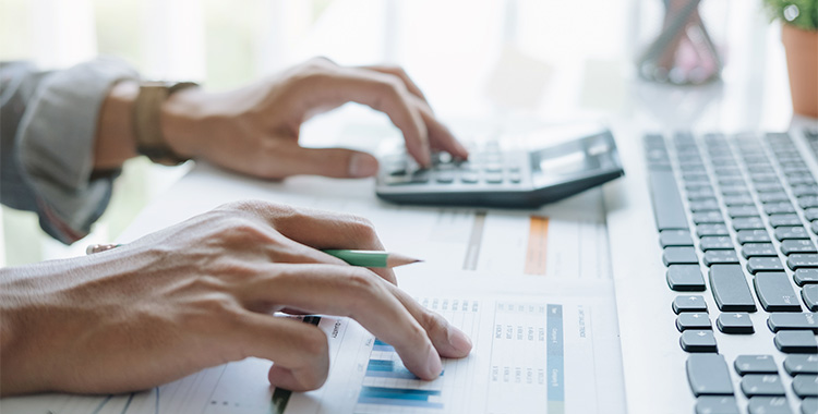 Uma fotografia focando em duas mãos de uma pessoa fazendo contas em uma calculadora e acompanhando um gráfico na mesa