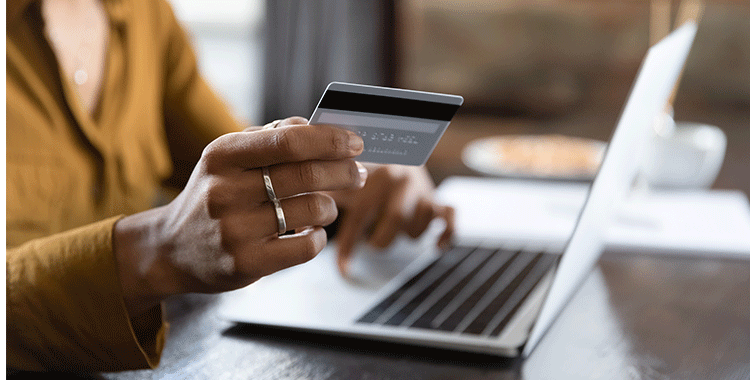 Fotografia da mão de uma mulher segurando o cartão de crédito no laptop, ao que parece estar realizando uma compra online