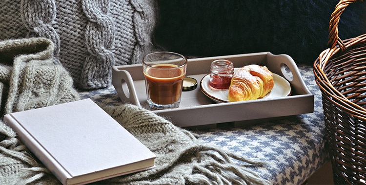 Uma fotografia de uma bandeja com xícara de café, um croissant  e um livro