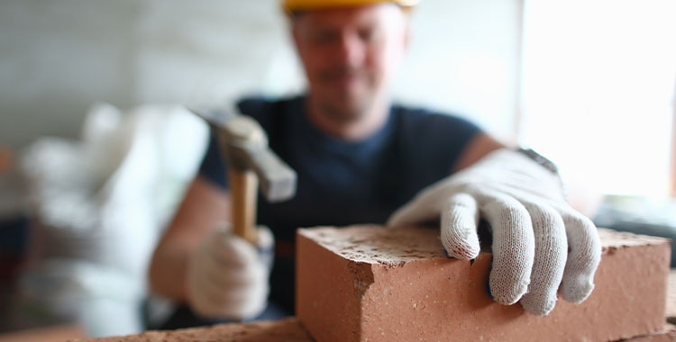 Uma foto de um homem segurando um tijolo e martelo referente a construção civil